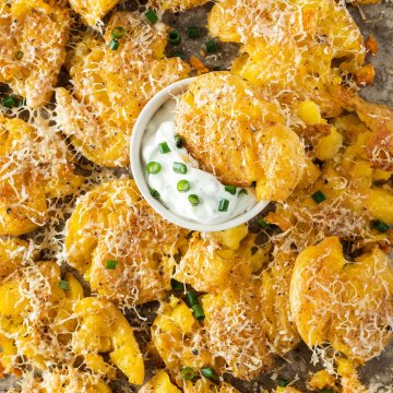 Nahaufnahme von mit Parmesan &uuml;berbackene Quetschkartoffeln mit einer kleinen Sourcream-Sch&uuml;ssel in der Mitte des Blechs.