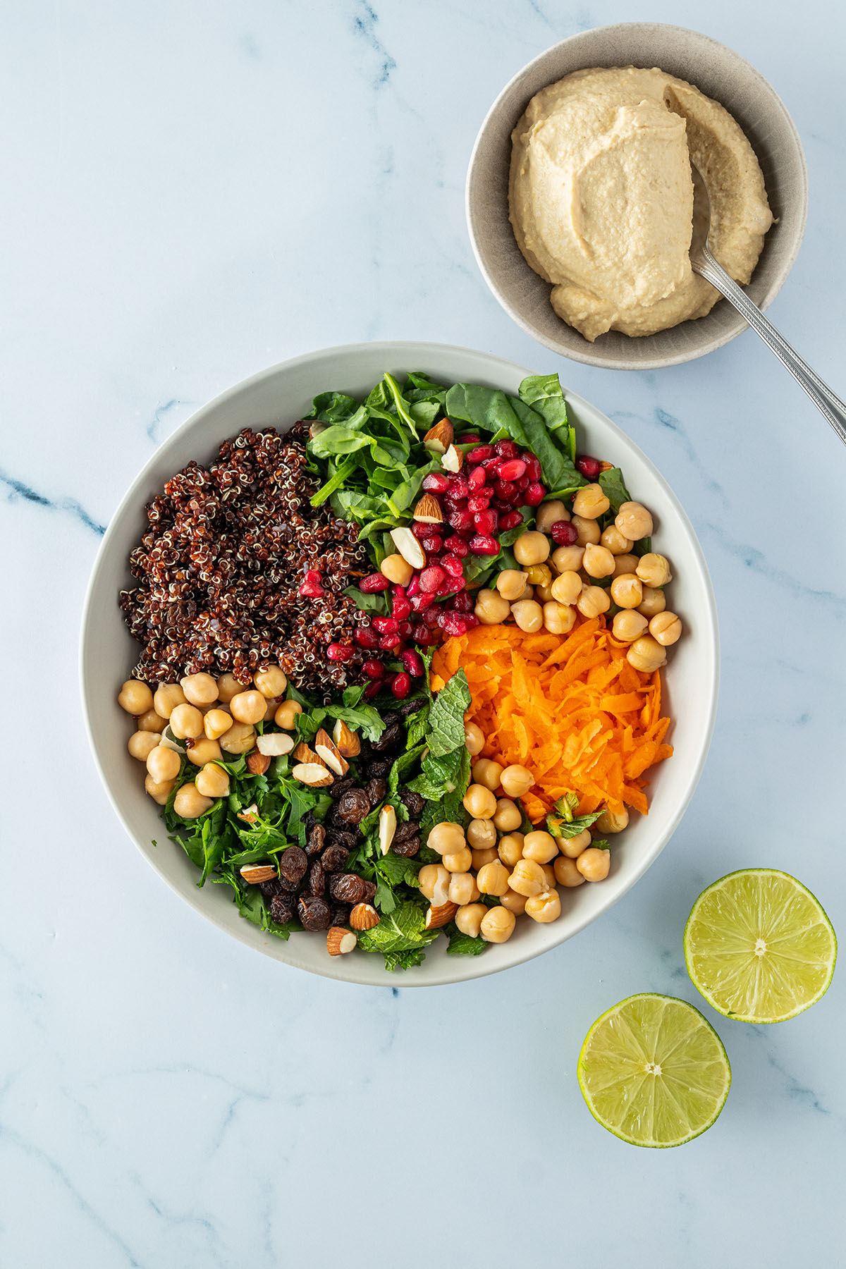 Alle Zutaten für den Quinoa Salat auf einem Teller, daneben eine Schüssel mit Hummus und eine halbierte Limette.