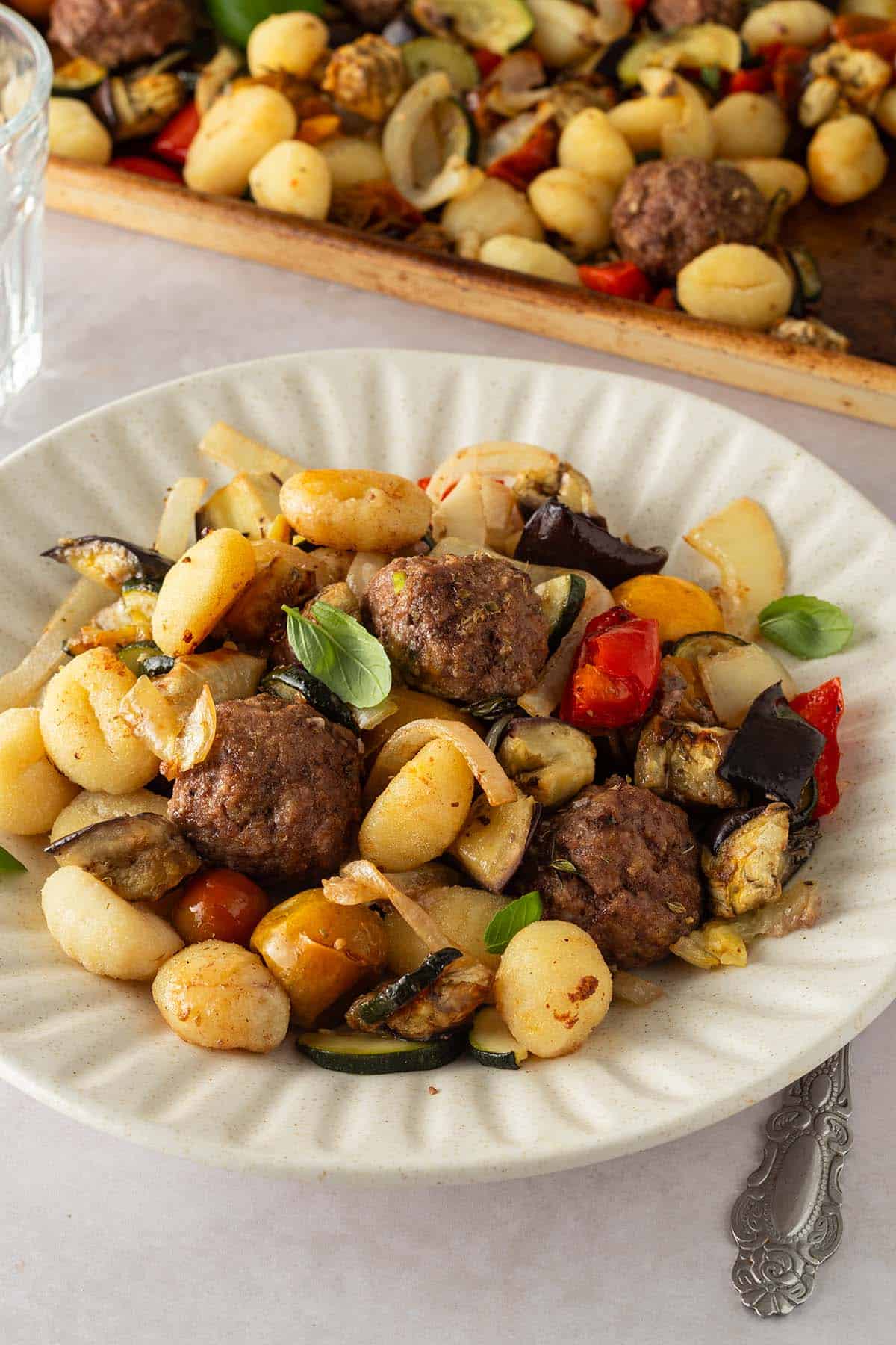 Eine Portion Ofen Ratatouille mit Gnocchi und Hackb&auml;llchen auf einem Teller, im Hintergrund das Blech.