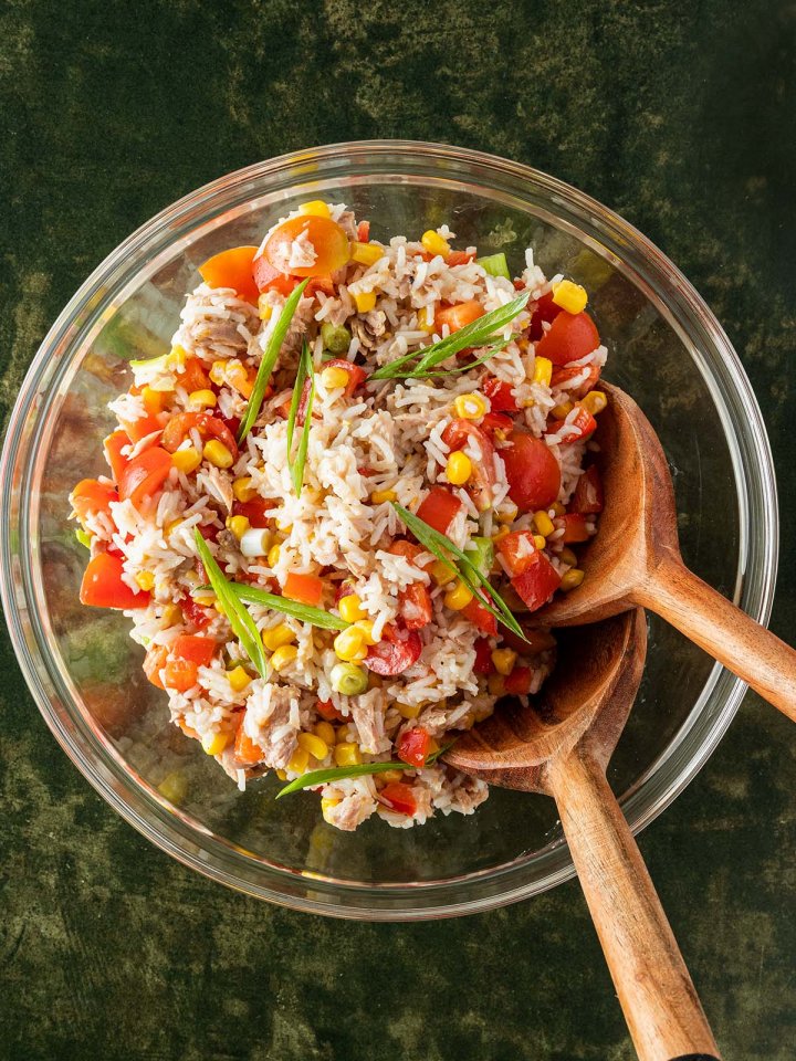 Eine gro&szlig;e Glassch&uuml;ssel mit einem Reissalat mit Thunfisch und Gem&uuml;se auf einem dunkelgr&uuml;nem Untergrund mit Salatbesteck aus Holz.
