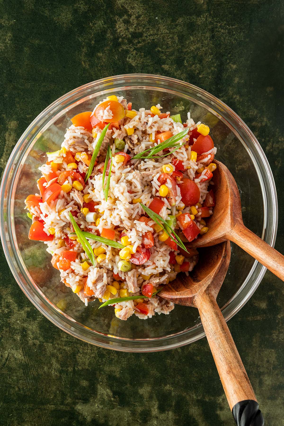 Eine gro&szlig;e Glassch&uuml;ssel mit einem Reissalat mit Thunfisch und Gem&uuml;se auf einem dunkelgr&uuml;nem Untergrund mit Salatbesteck aus Holz.
