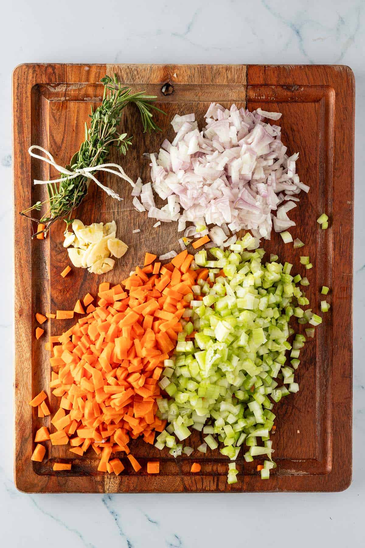 Fein gew&uuml;rfelte Karotten, Sellerie, Schalotten, in d&uuml;nne Scheiben geschnittener Knoblauch und zu einem B&uuml;ndel gebundene Kr&auml;uter auf einem Holzbrett.