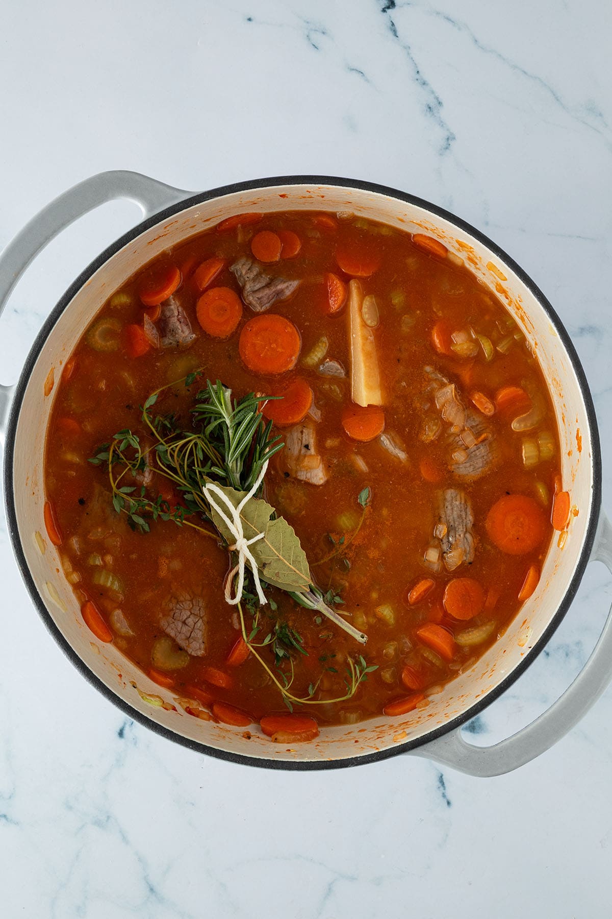 Rindfleischeintopf im Topf mit Kr&auml;uterb&uuml;ndel aus Rosmarin, Thymian und Lorbeerblatt sowie einem St&uuml;ck Parmesanrinde.