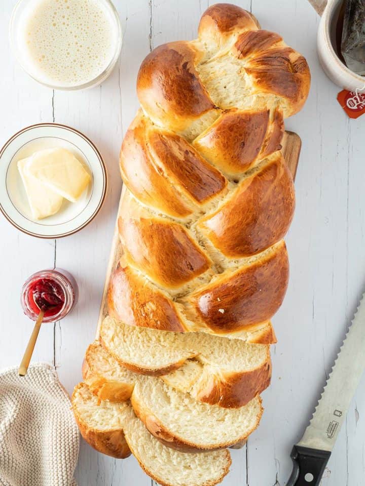 Ein wei&szlig;er Holztisch mit einem angeschnittenen Hefezopf, au&szlig;en herum Tassen, Butter, Marmelade und weitere Fr&uuml;hst&uuml;cksutensilien.