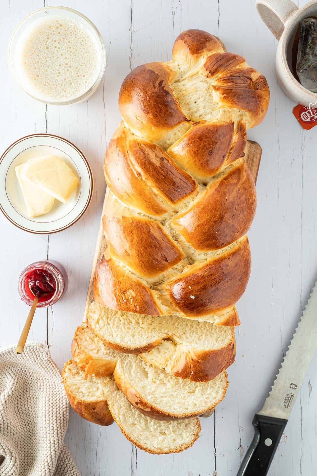 Ein wei&szlig;er Holztisch mit einem angeschnittenen Hefezopf, au&szlig;en herum Tassen, Butter, Marmelade und weitere Fr&uuml;hst&uuml;cksutensilien.