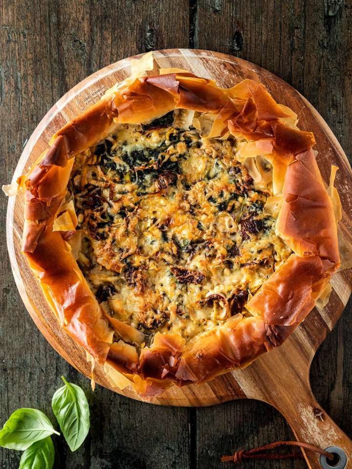 Gem&uuml;se-Quiche mit Filo-Teig auf einem Holzbrett auf einem rustikalen Holzuntergrund, Basilikumbl&auml;tter im Bildrand.