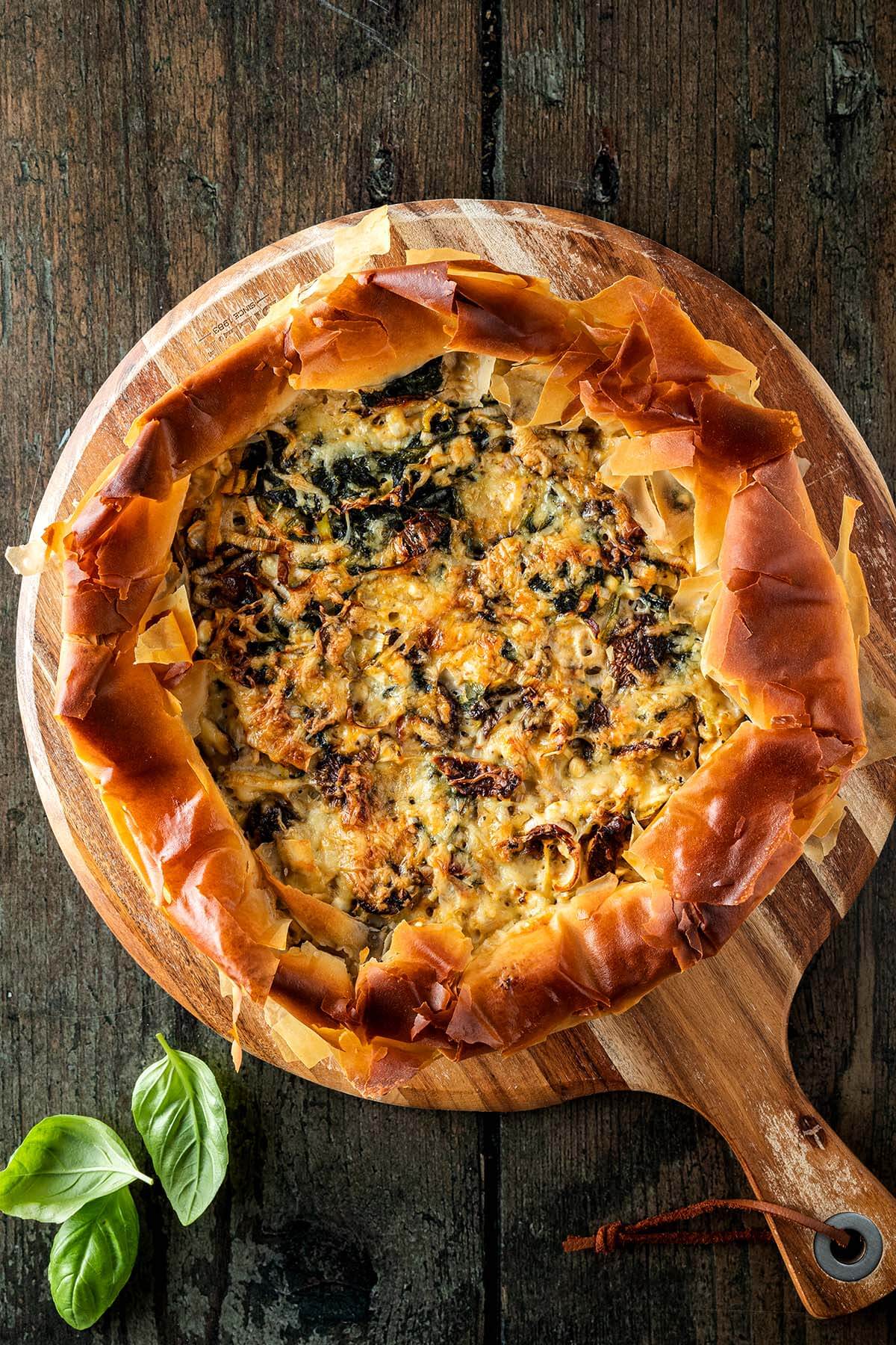 Gem&uuml;se-Quiche mit Filo-Teig auf einem Holzbrett auf einem rustikalen Holzuntergrund, Basilikumbl&auml;tter im Bildrand.