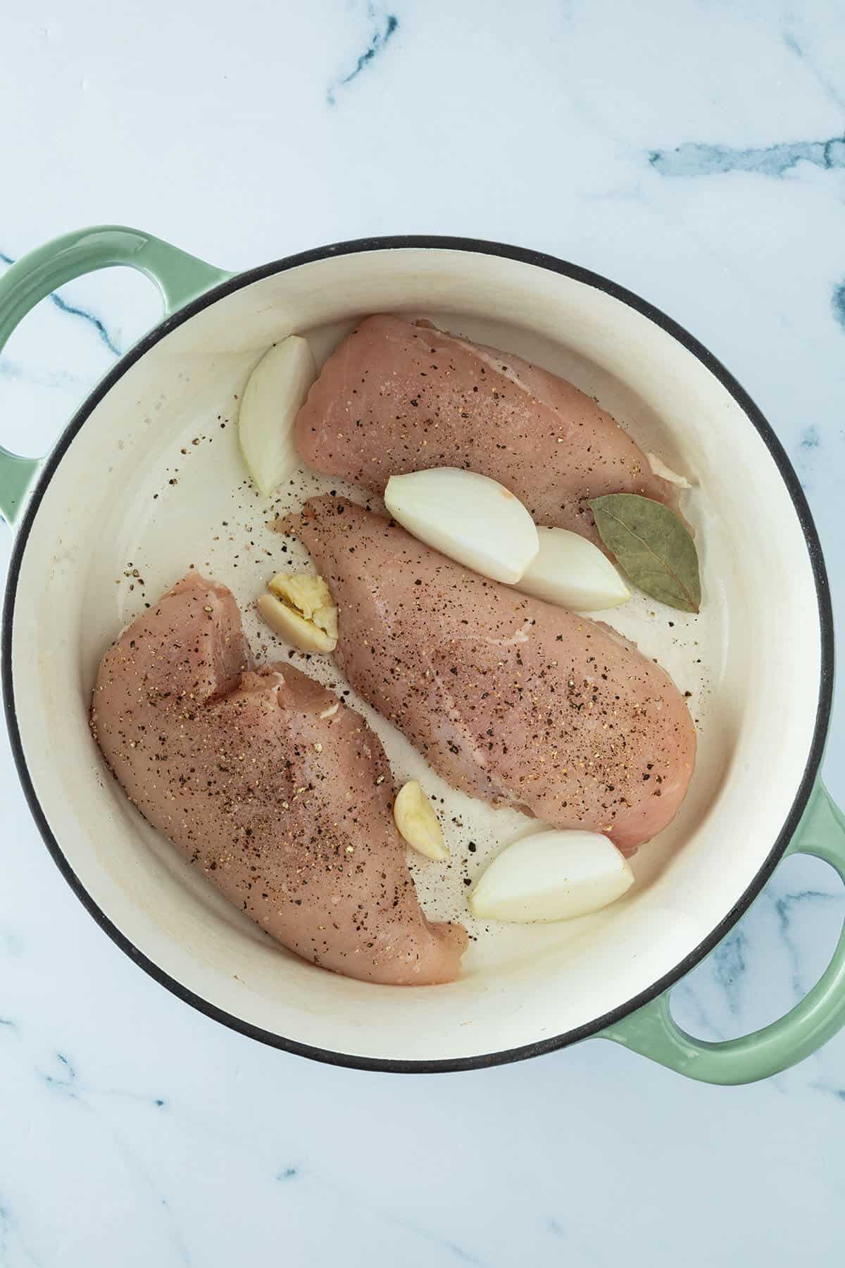H&auml;hnchenbrustfilets in einem Topf mit Knoblauch, Zwiebeln und einem Lorbeerblatt.