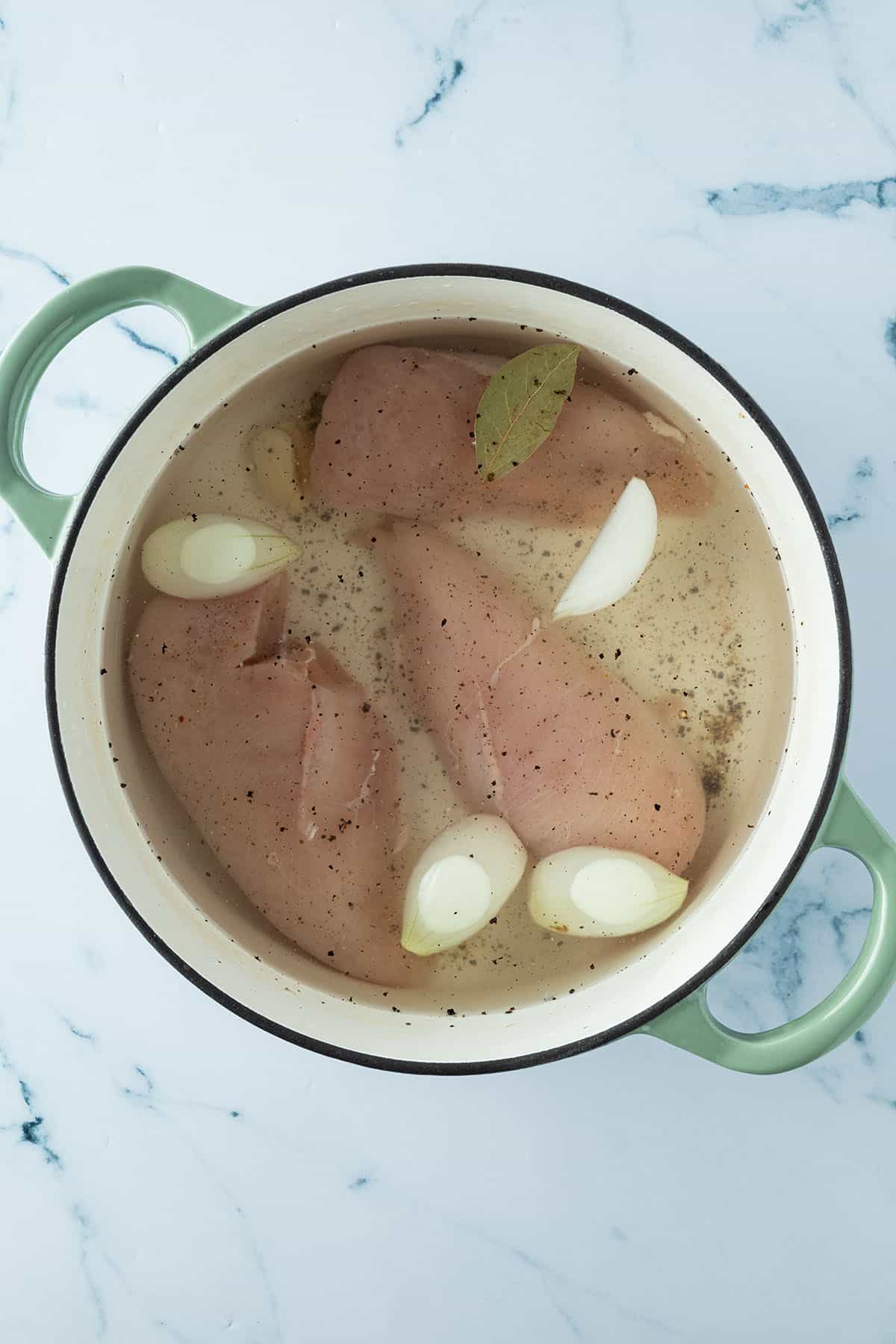 Rohe H&auml;hnchenfilets in einem Topf mit Gew&uuml;rzen, Zwiebeln und Knoblauch, die mit Wasser bedeckt sind.