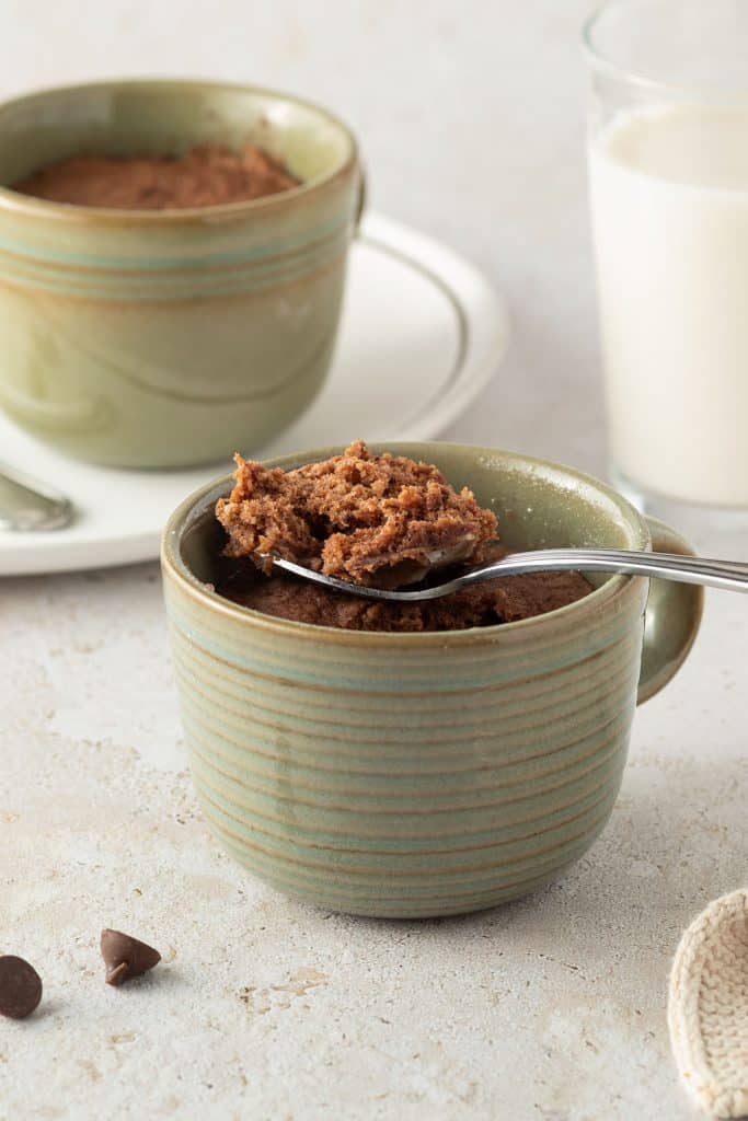 Schoko-Tassenkuchen in einer Kaffeetasse, zwei Kuchen und ein Glas Milch im Hintergrund.