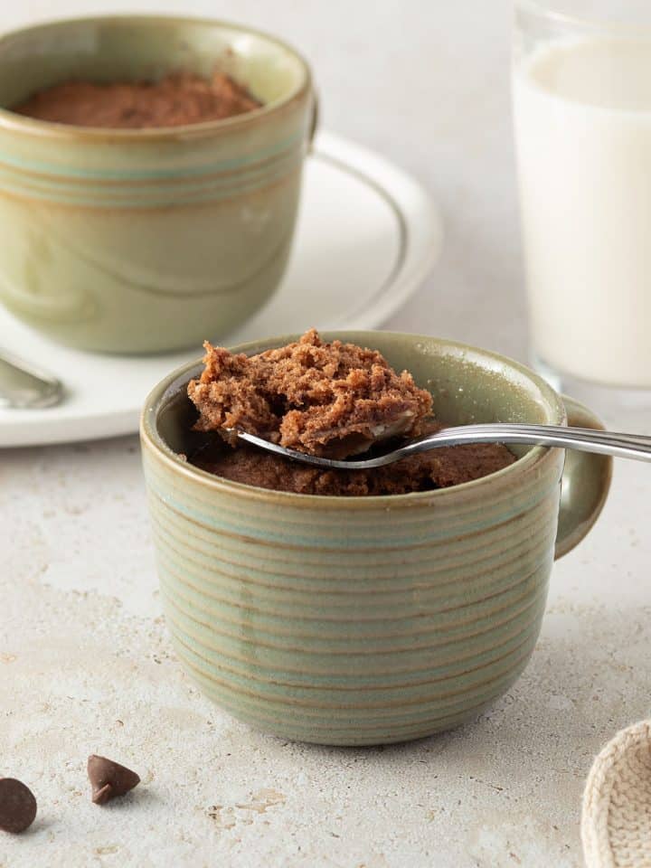Schoko-Tassenkuchen in einer Kaffeetasse, zwei Kuchen und ein Glas Milch im Hintergrund.