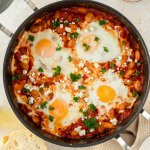 Vogelperspektive auf eine Pfanne mit Shakshuka, das mit frischer Petersilie und Kr&auml;utern serviert wird, Deko im Bildrand.