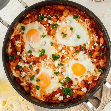 Vogelperspektive auf eine Pfanne mit Shakshuka, das mit frischer Petersilie und Kr&auml;utern serviert wird, Deko im Bildrand.