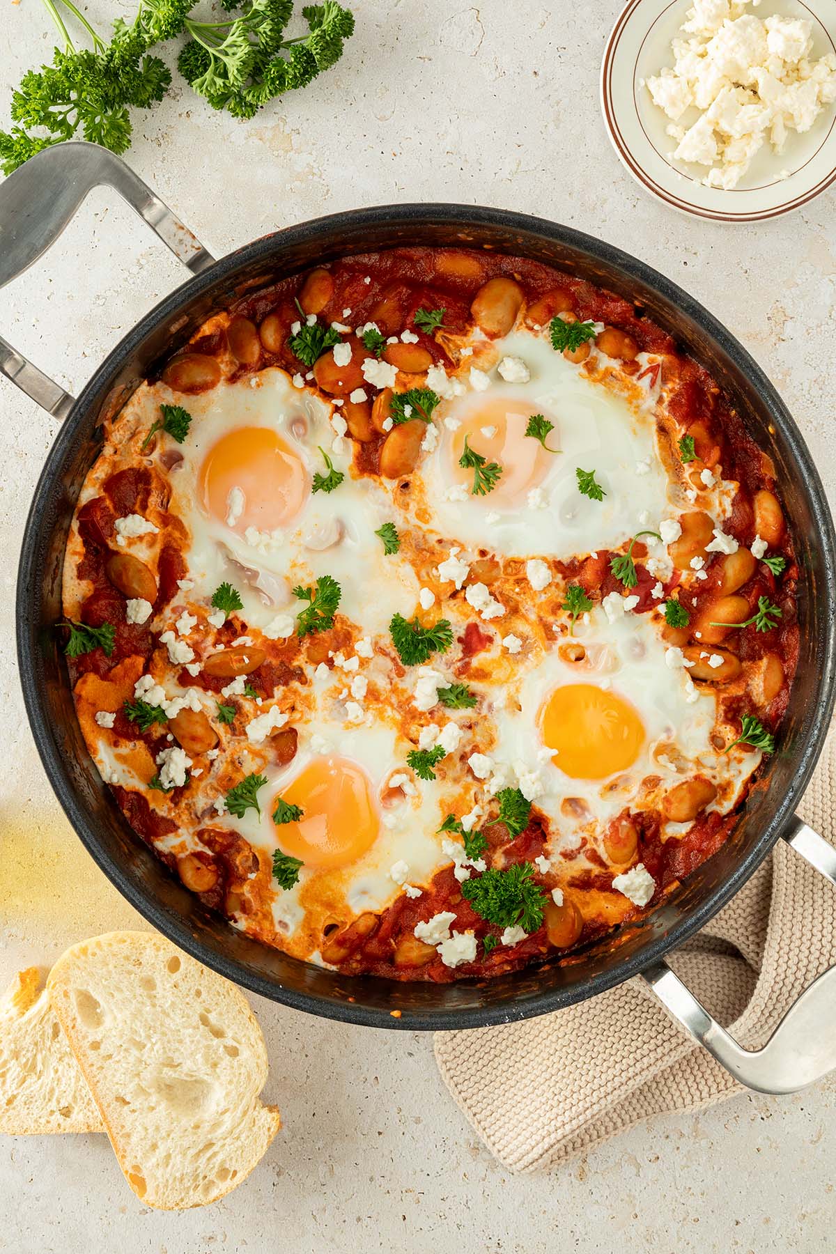 Vogelperspektive auf eine Pfanne mit Shakshuka, das mit frischer Petersilie und Kr&auml;utern serviert wird, Deko im Bildrand.