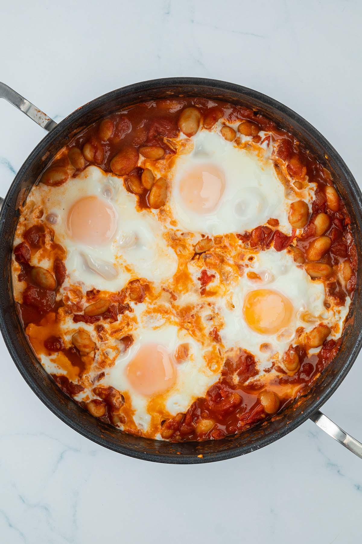 Vogelperspektive auf eine Pfanne mit vier, in einer Tomatensauce mit Bohnen pochierte Eier.