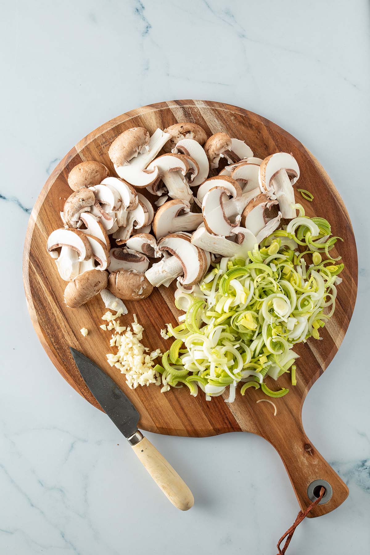 Dünne Lauchringe, Champignon-Scheiben und fein gewürfelter Knoblauch auf einem Holzbrett mit einem Küchenmesser daneben.