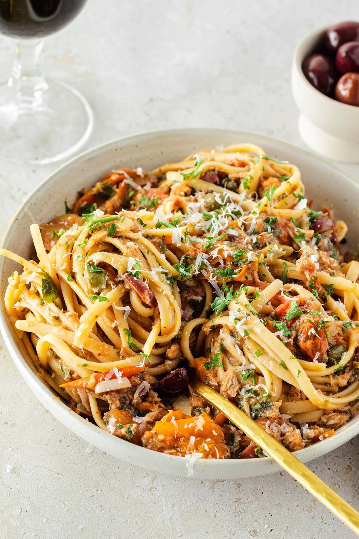 Ein Teller Spaghetti alla Puttanesca mit frischen Kr&auml;utern und Parmesan, im Hintergrund Oliven und ein Glas Wein.