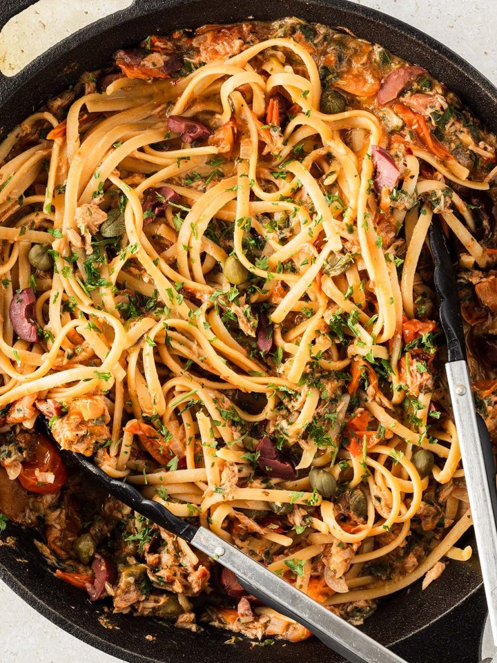 Nahaufnahme von Spaghetti Puttanesca mit Thunfisch in einer schwarzen Pfanne mit einer K&uuml;chenzange.