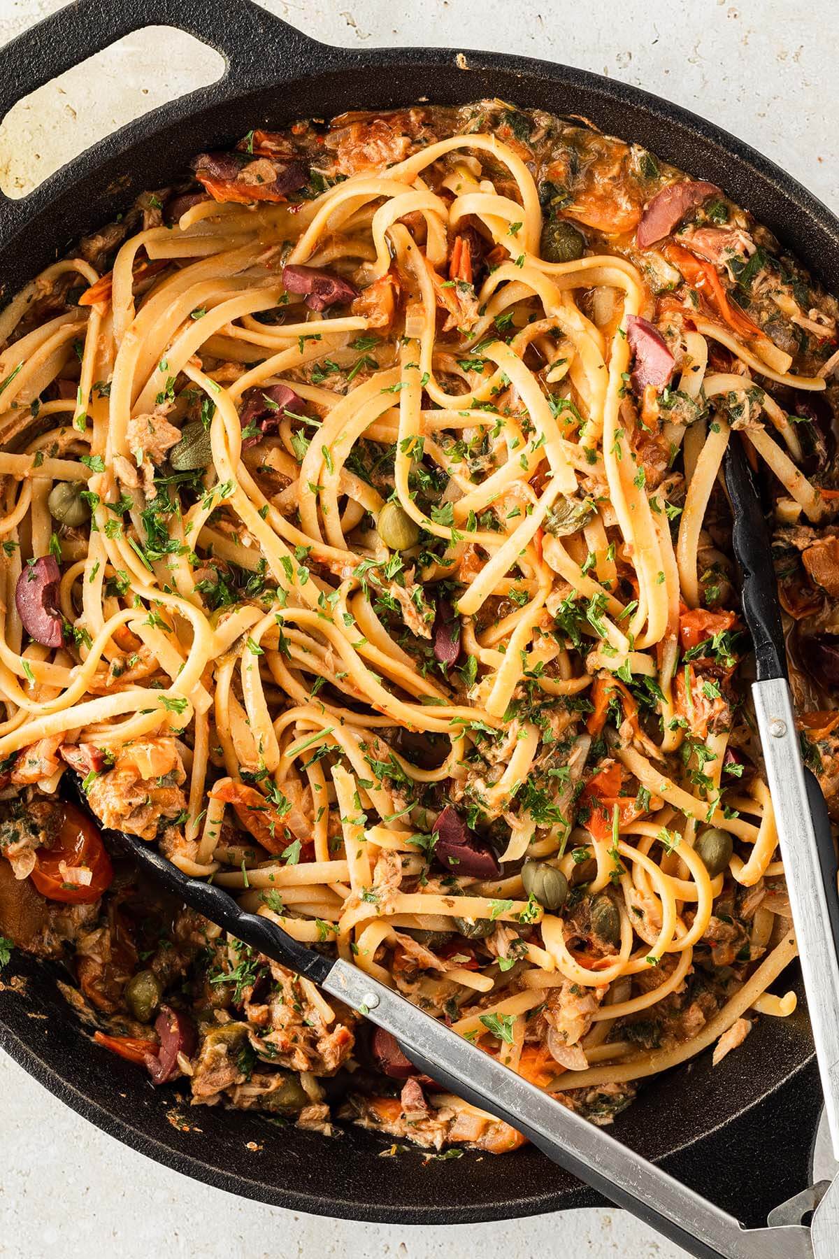 Nahaufnahme von Spaghetti Puttanesca mit Thunfisch in einer schwarzen Pfanne mit einer K&uuml;chenzange.