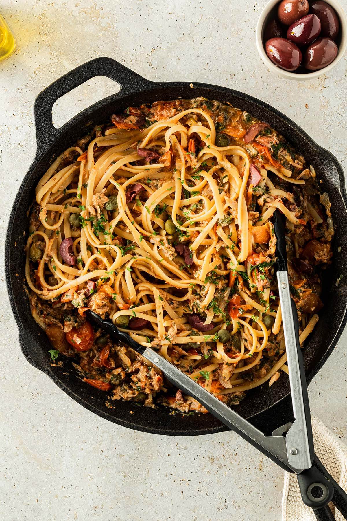 Spaghetti Puttanesca serviert in einer Gusseisenpfanne, Deko im Bildrand.