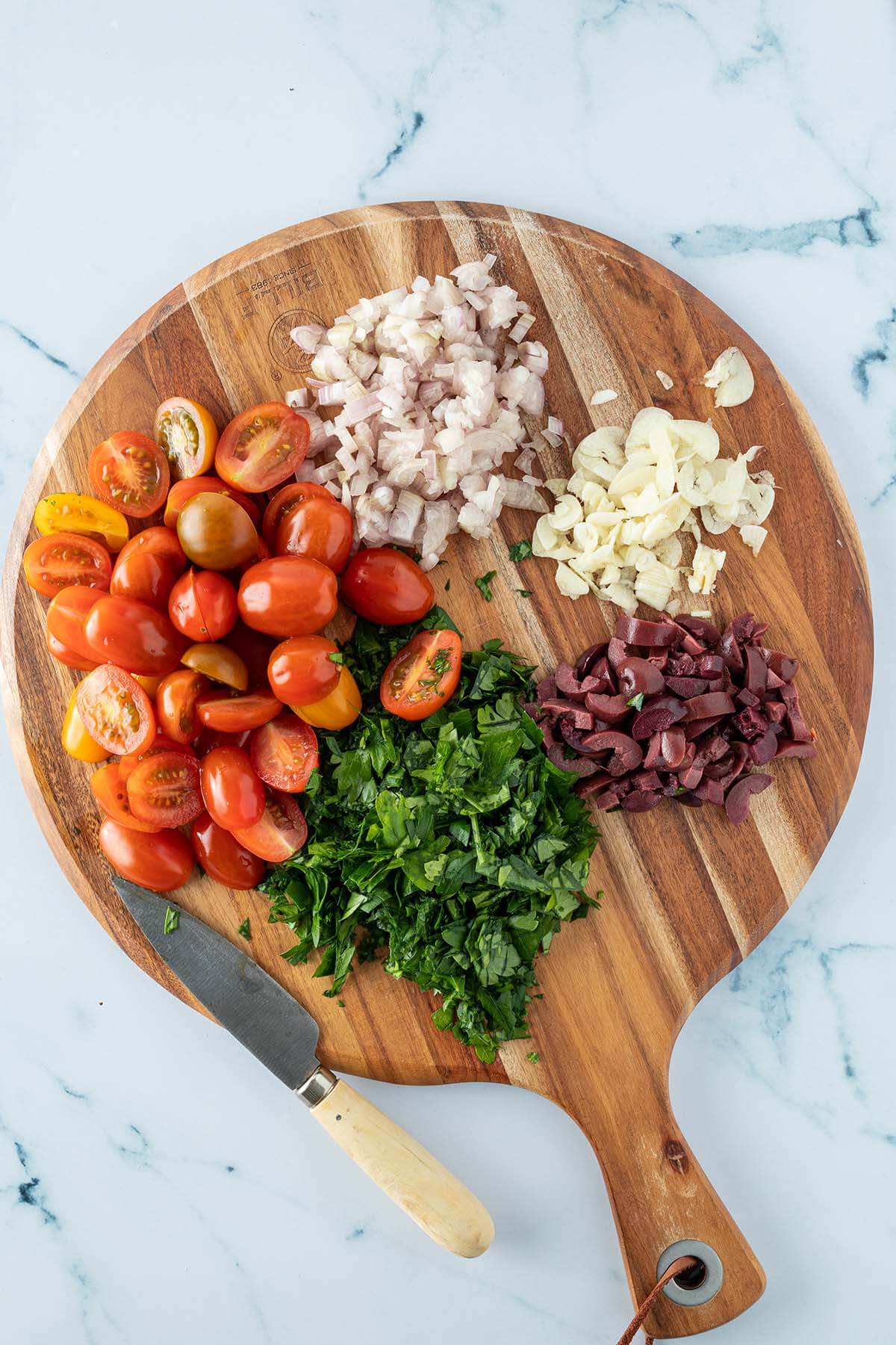 Rundes Holzbrett mit halbierten Tomaten, gehackter Petersilie, gew&uuml;rfelten Schalotten, Knoblauchscheiben und kleingeschnittenen Oliven.