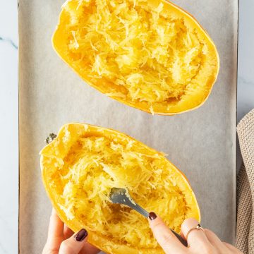 Zwei gegarte Spaghettik&uuml;rbis-H&auml;lften auf einem Blech, wo zwei H&auml;nde das Fruchtfleisch von einer H&auml;lfte mit einer Gabel auskratzen.