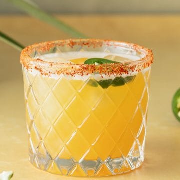Spicy Margarita Cocktail in einem Glas mit Chili-Salzrand und einer Jalape&ntilde;o-Scheibe auf einem gelben Untergrund.