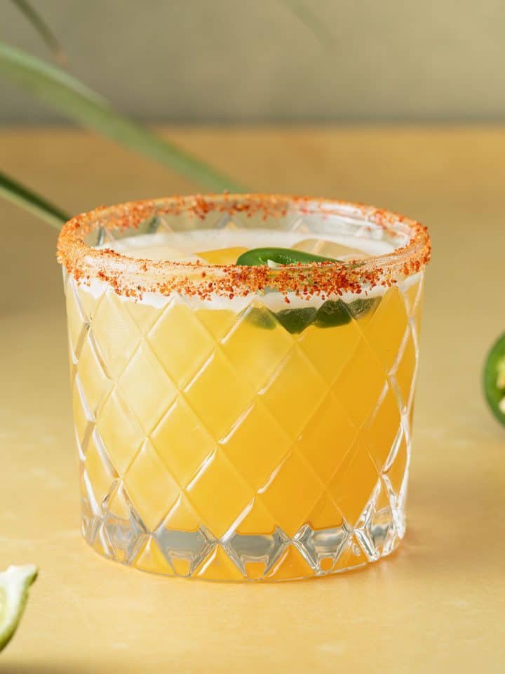 Spicy Margarita Cocktail in einem Glas mit Chili-Salzrand und einer Jalape&ntilde;o-Scheibe auf einem gelben Untergrund.