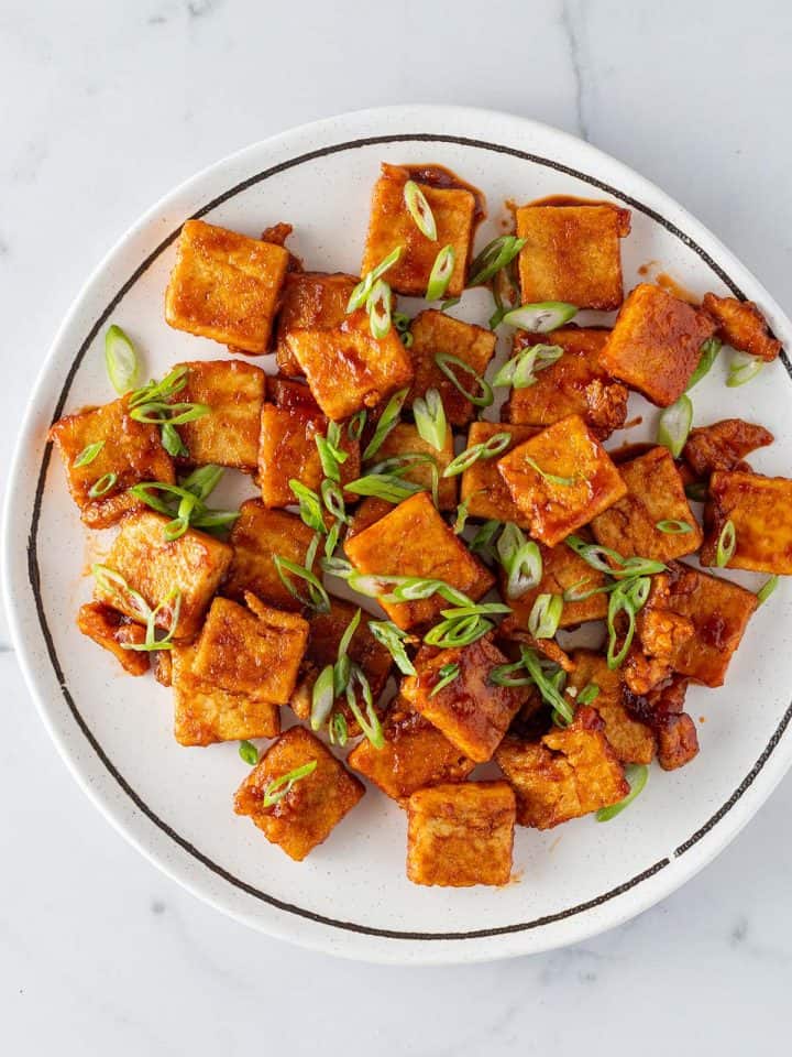 Knusprige Tofu-St&uuml;cke mit einer Sticky s&uuml;&szlig;-sauer Sauce auf einem Teller mit Fr&uuml;hlingszwiebeln.