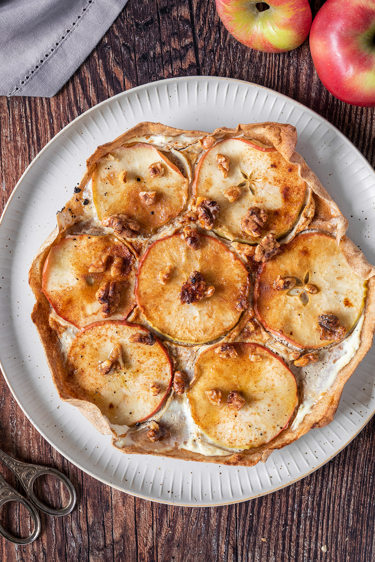 S&uuml;&szlig;er Flammkuchen mit in Scheiben geschnittenen &Auml;pfeln, Ricotta und Waln&uuml;ssen belegt und mit etwas Eis serviert vor einem h&ouml;lzernem Hintergrund.