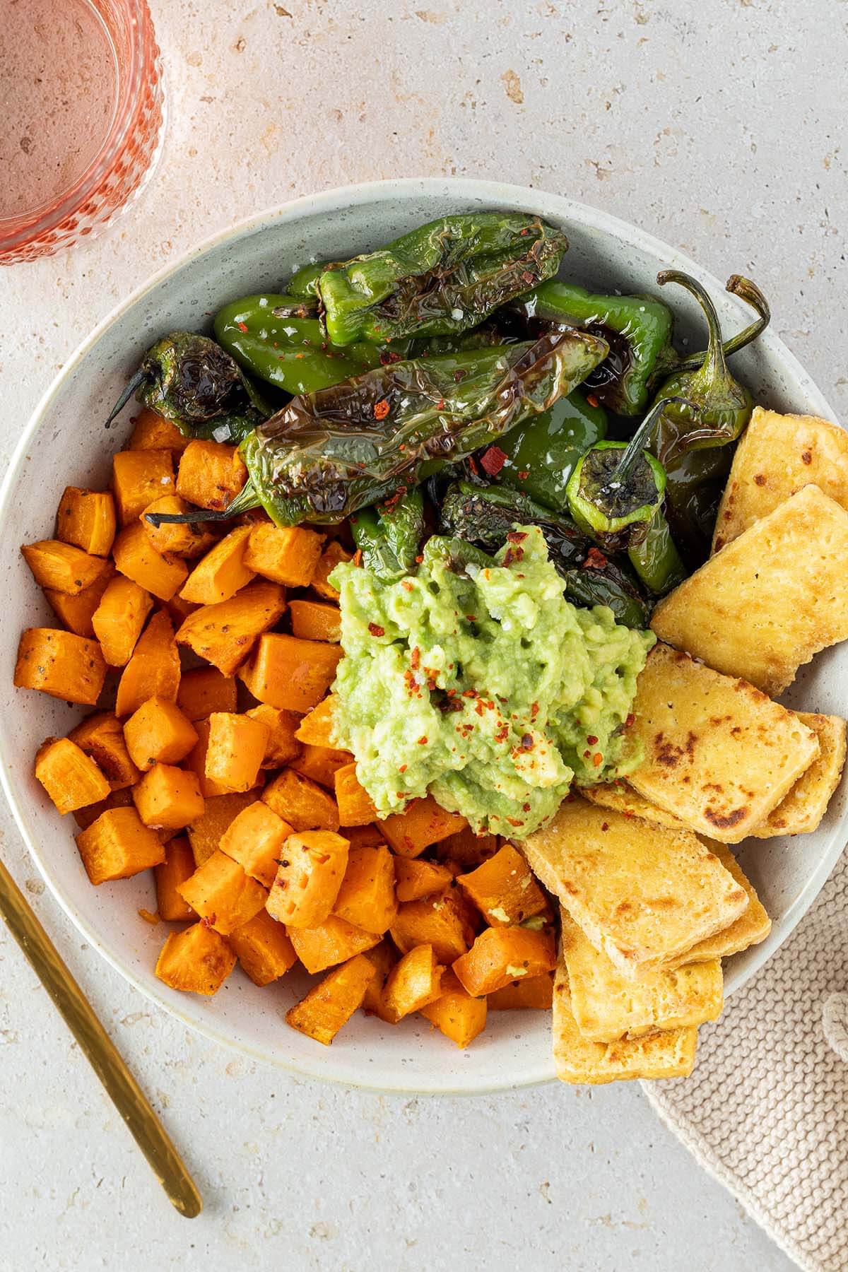 Sch&uuml;ssel mit S&uuml;&szlig;kartoffelw&uuml;rfel, angebratenen Pimientos, knusprigen Tofu-Scheiben und Guacamole in der Mitte.