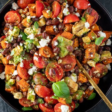 S&uuml;&szlig;kartoffelsalat mit Avocado, Bohnen, Tomaten und einem gr&uuml;nen Dressing auf einem schwarzen Teller auf dunklem Untergrund.