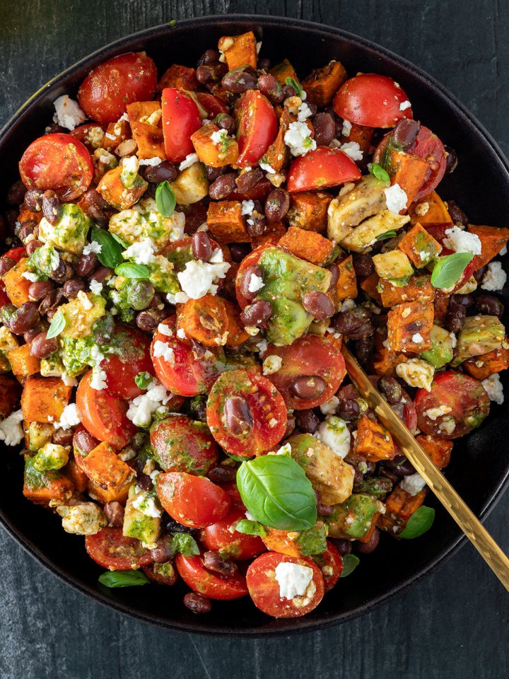 S&uuml;&szlig;kartoffelsalat mit Avocado, Bohnen, Tomaten und einem gr&uuml;nen Dressing auf einem schwarzen Teller auf dunklem Untergrund.