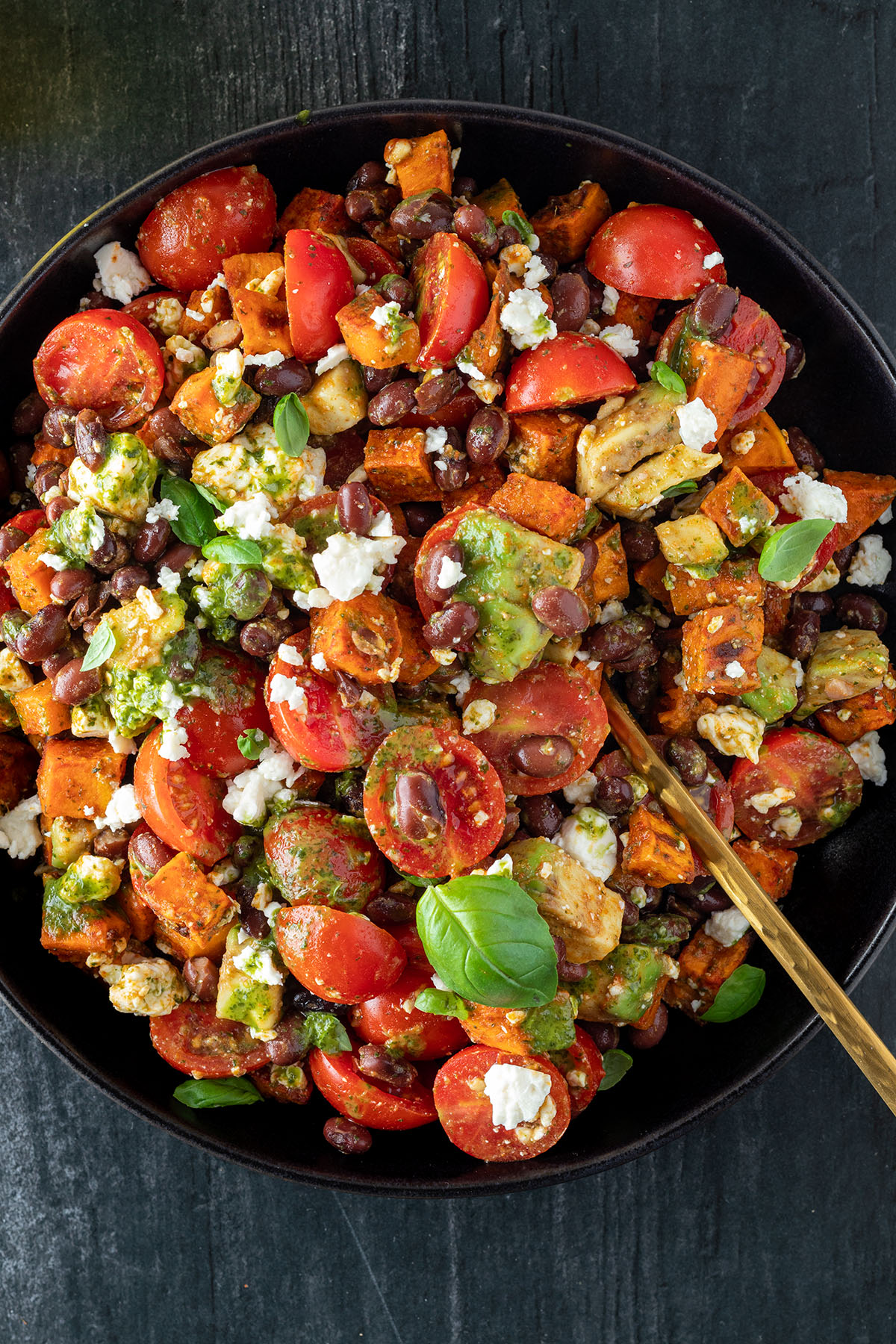 S&uuml;&szlig;kartoffelsalat mit Avocado, Bohnen, Tomaten und einem gr&uuml;nen Dressing auf einem schwarzen Teller auf dunklem Untergrund.