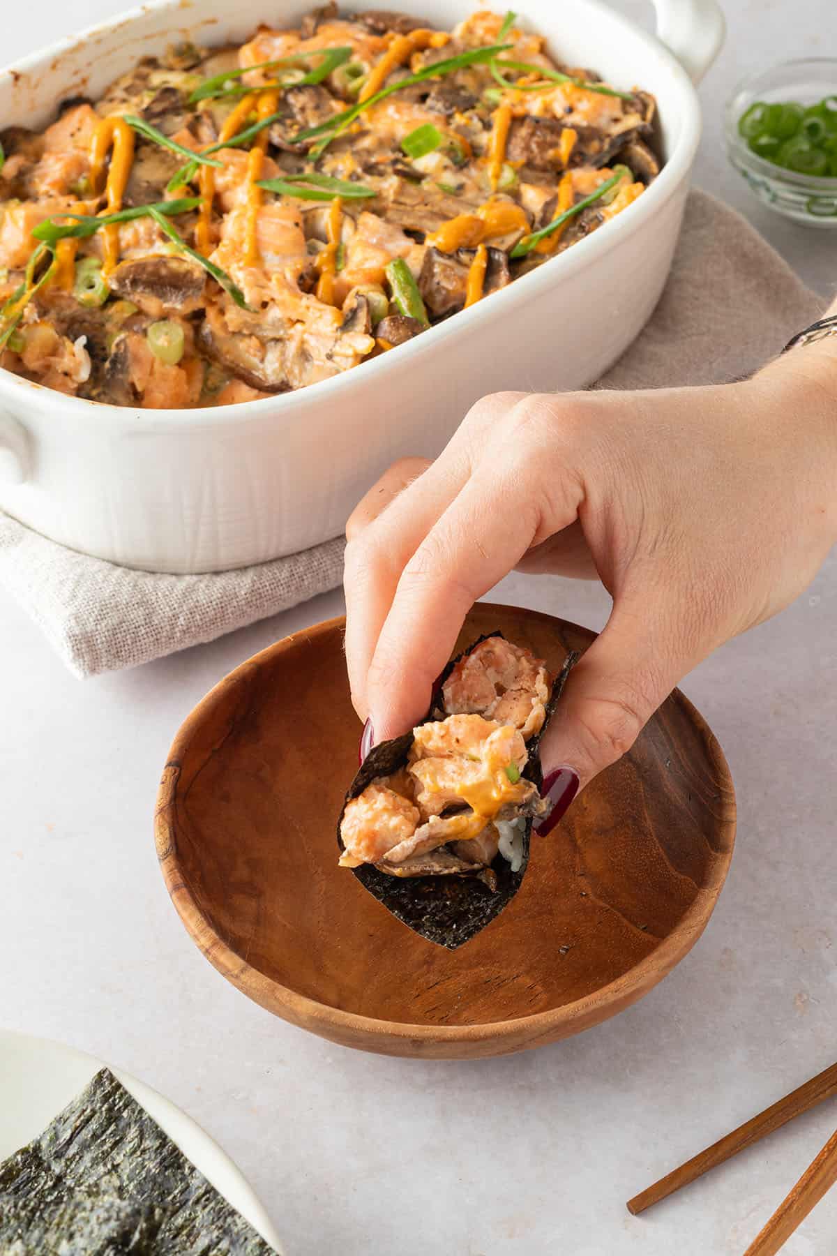 Eine Hand h&auml;lt ein kleines St&uuml;ck Nori-Blatt, das mit etwas Sushi-Auflauf gef&uuml;llt ist. Im Hintergrund steht der Sushi-Bake.