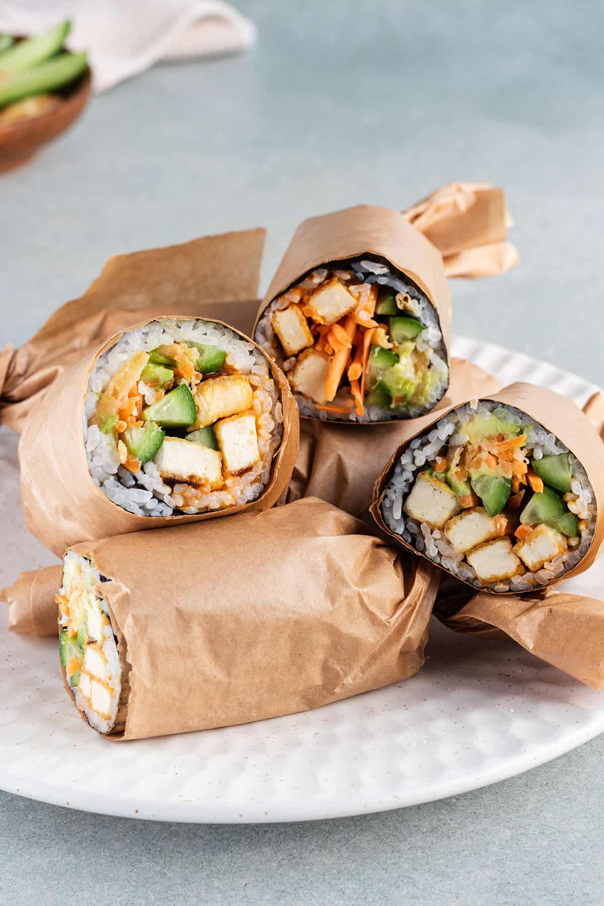 Mehrere, halbierte und in Papier eingewickelte Sushi Burritos mit Tofu auf einem Teller.