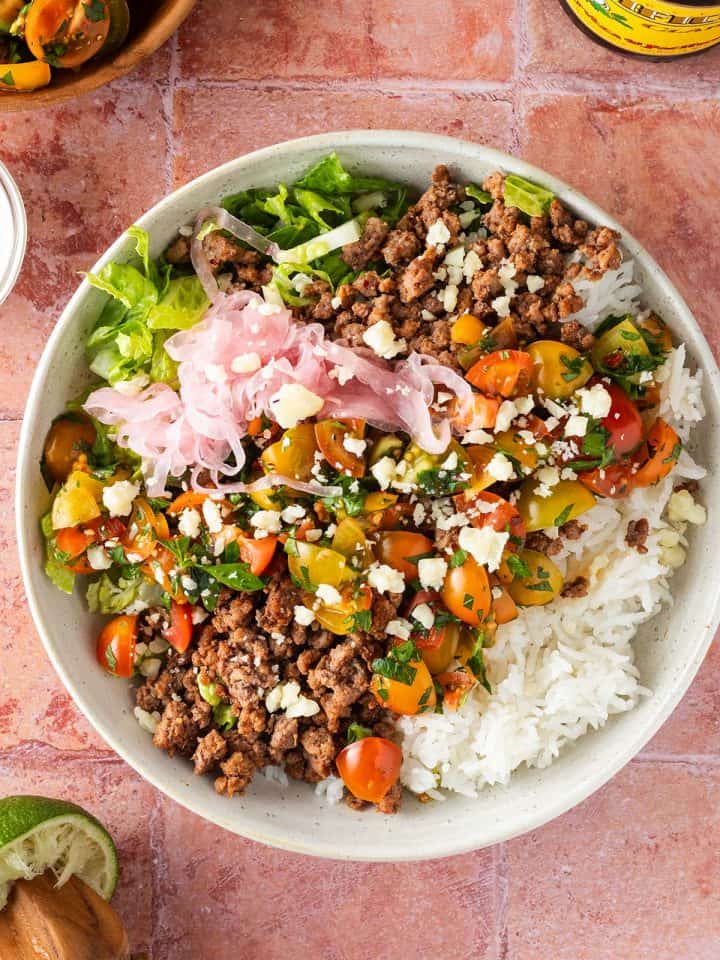 Taco Bowl mit Hackfleisch, Salsa, Reis und Salat auf einem roten Flie&szlig;enuntergrund, Deko im Bildrand.