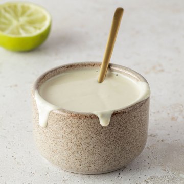 Kleine Sch&uuml;ssel mit einer cremigen, hellen Tahini Sauce und einem goldenen L&ouml;ffeln, eine halbe Limette im Hintergrund.