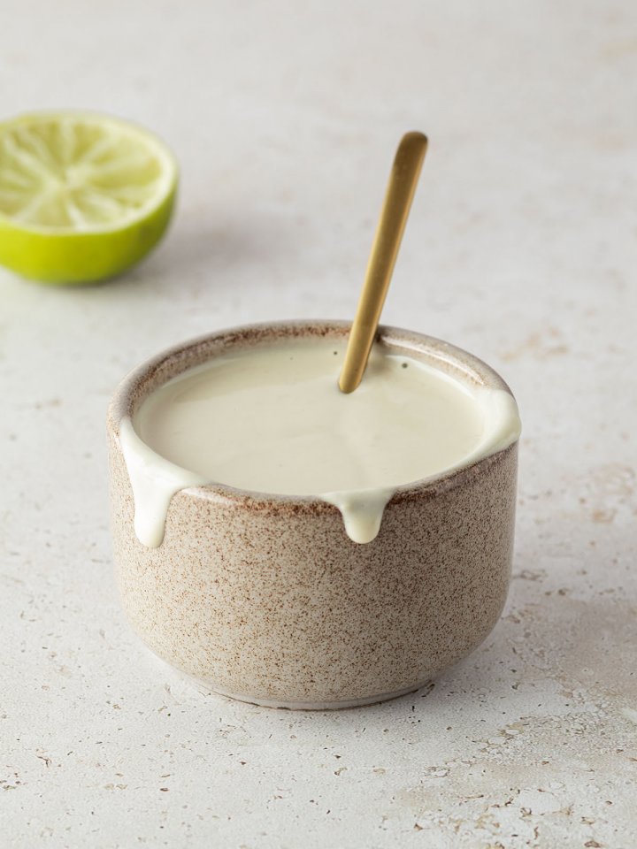 Kleine Sch&uuml;ssel mit einer cremigen, hellen Tahini Sauce und einem goldenen L&ouml;ffeln, eine halbe Limette im Hintergrund.