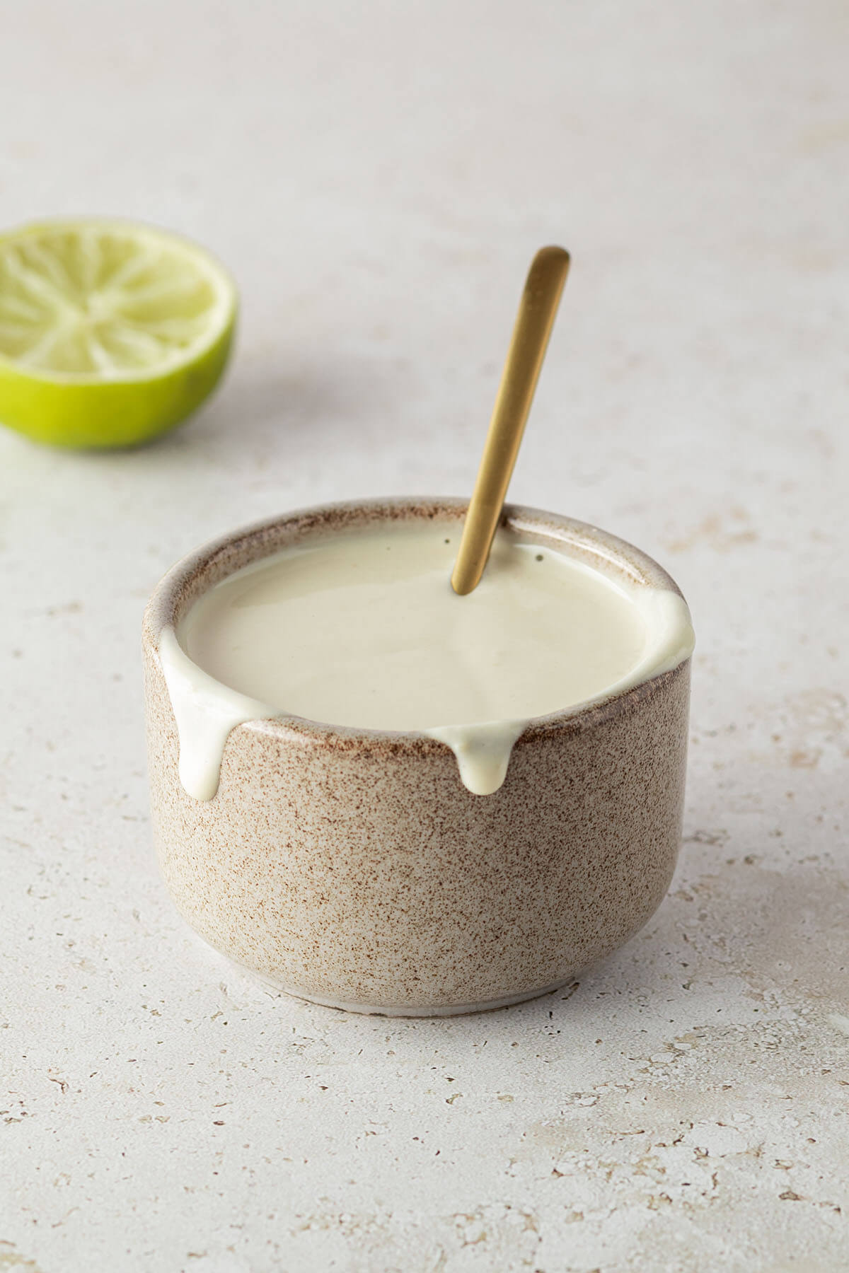Kleine Sch&uuml;ssel mit einer cremigen, hellen Tahini Sauce und einem goldenen L&ouml;ffeln, eine halbe Limette im Hintergrund.