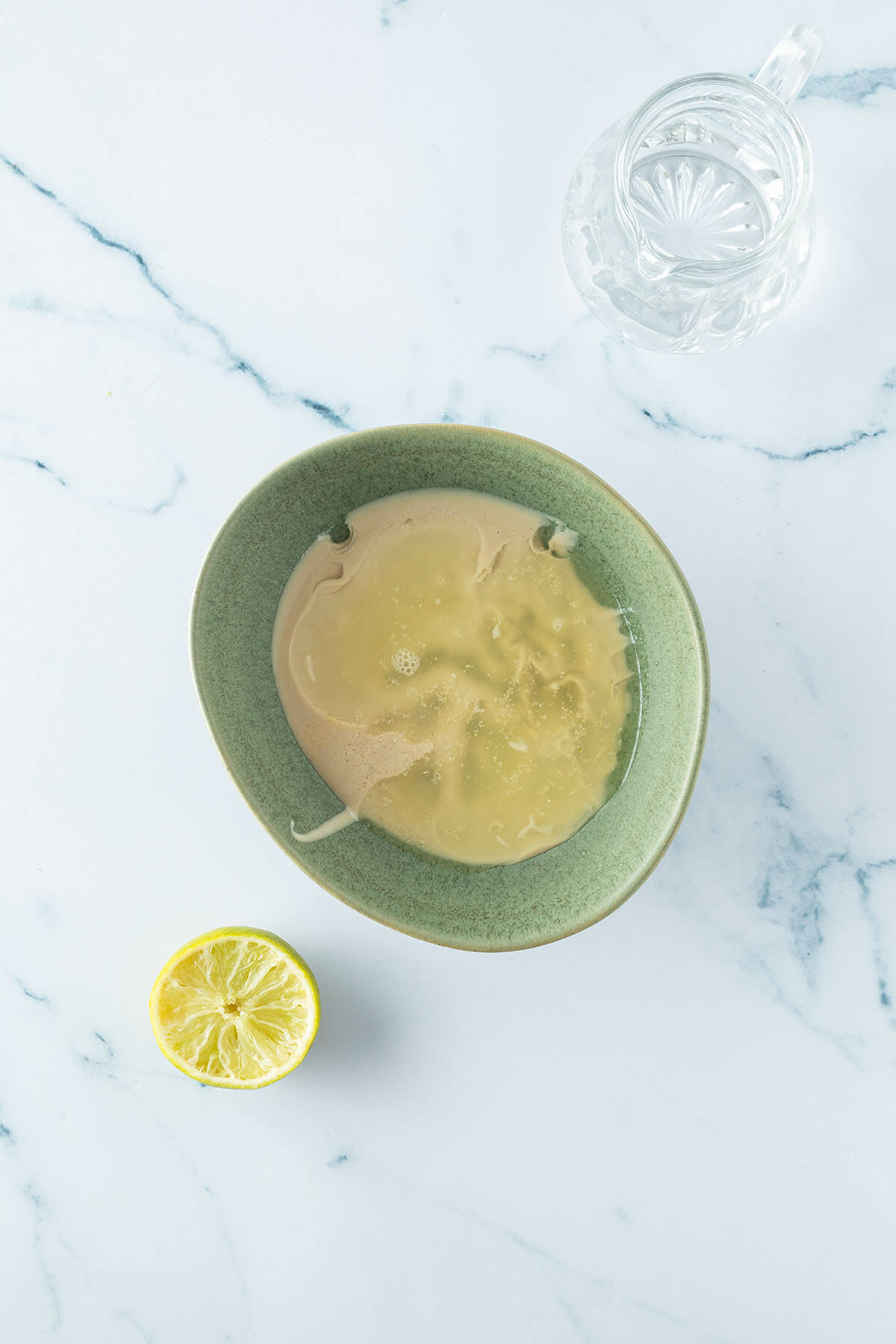 Einzelne Zutaten f&uuml;r die Tahini Sauce unverr&uuml;hrt in einer gr&uuml;nen Sch&uuml;ssel mit einer Karaffe Wasser und einer ausgepressten Limette daneben.