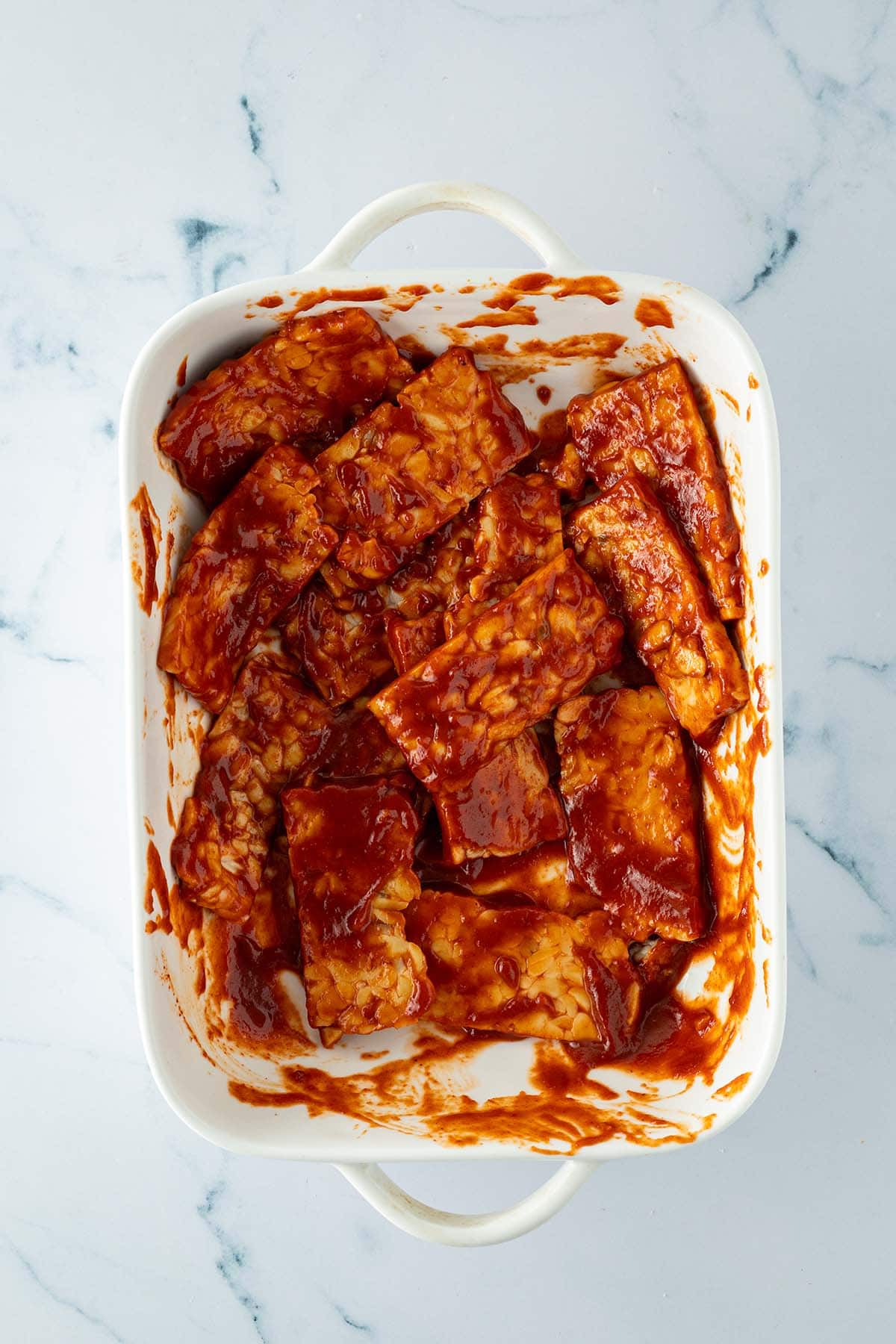 In Scheiben geschnittener Tempeh wird in einer Auflaufform mit einer dickfl&uuml;ssigen BBQ Sauce mariniert.