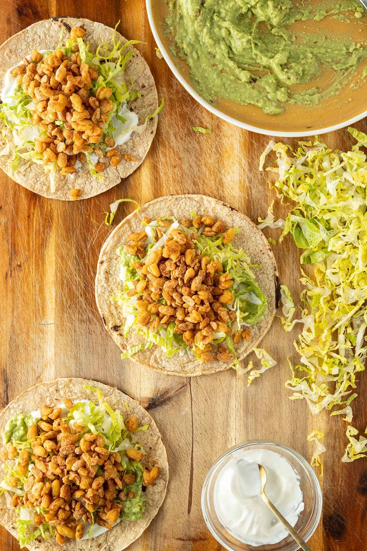 Drei Tacos auf einem Holzbrett, gef&uuml;llt mit Tempeh, Salat, Guacamole und Schmand, daneben Zutaten in Sch&uuml;sseln.