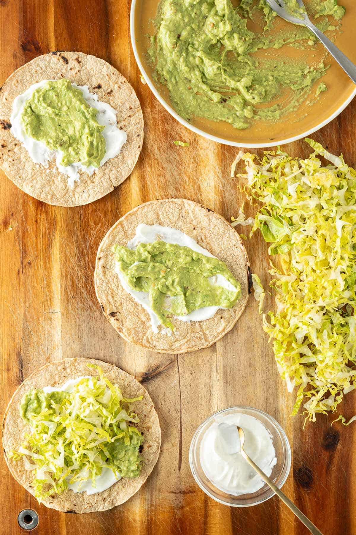 Drei Tacos auf einem Holzbrett getoppt mit Schmand und Guacamole, daneben Sch&uuml;sseln mit Zutaten.