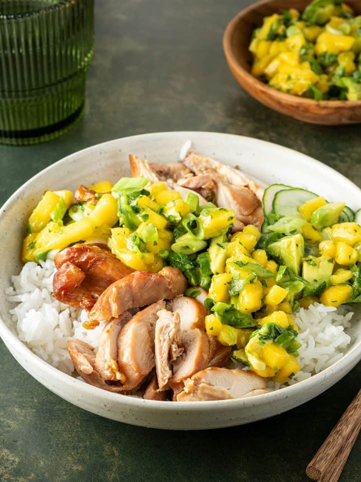 Teriyaki Bowl mit H&auml;hnchen, Reis und Mango-Salsa auf einem Tisch, ein Glas und eine Sch&uuml;ssel mit Salsa im Hintergrund.