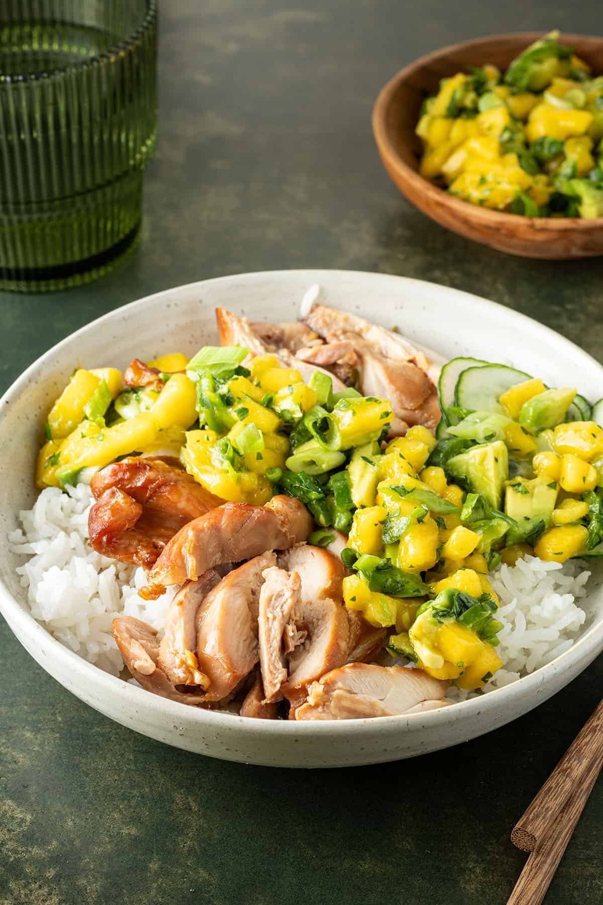 Teriyaki Bowl mit H&auml;hnchen, Reis und Mango-Salsa auf einem Tisch, ein Glas und eine Sch&uuml;ssel mit Salsa im Hintergrund.