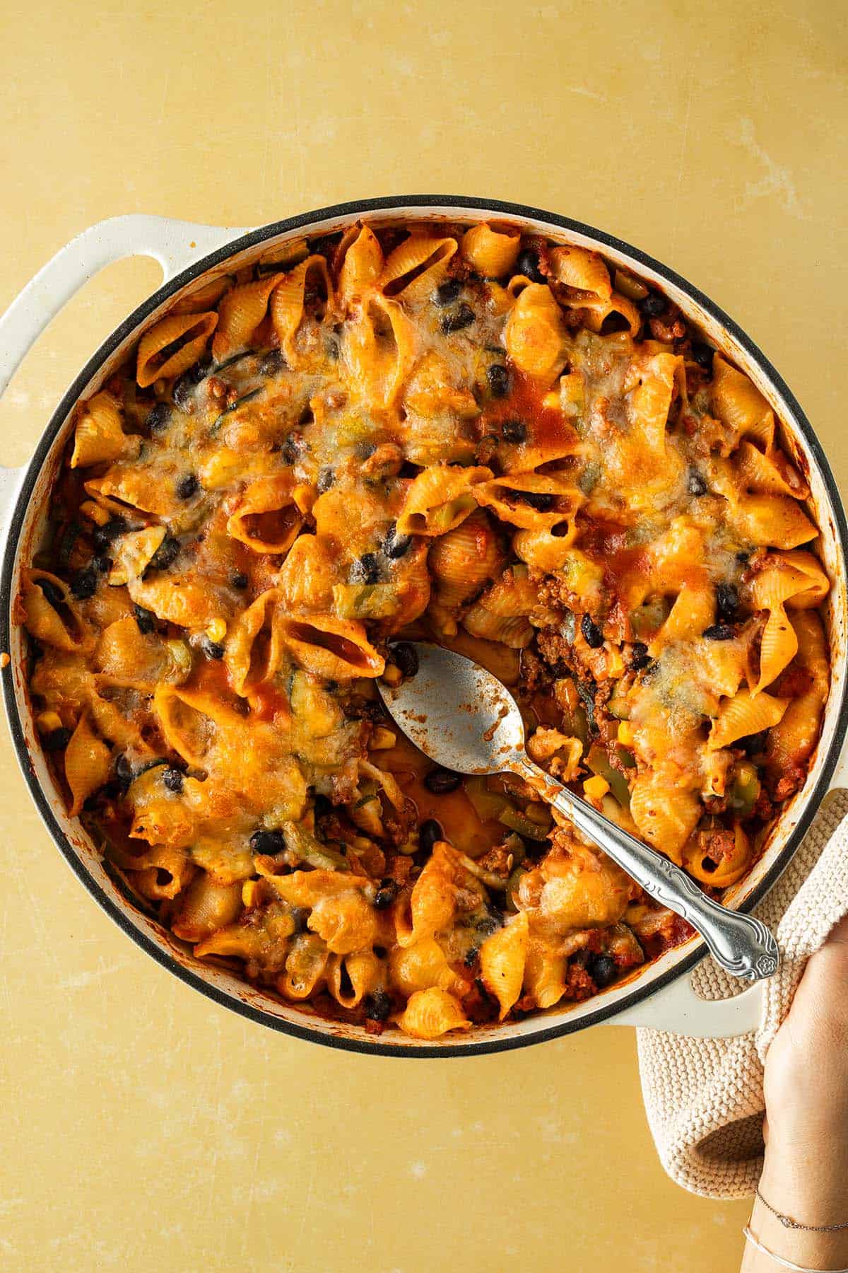 Auflaufform mit &uuml;berbackenem Tex-Mex Nudelauflauf aus Conchiglie, Hackfleisch, schwarzen Bohnen und geschmolzenem K&auml;se, ein L&ouml;ffel hebt eine Portion heraus.