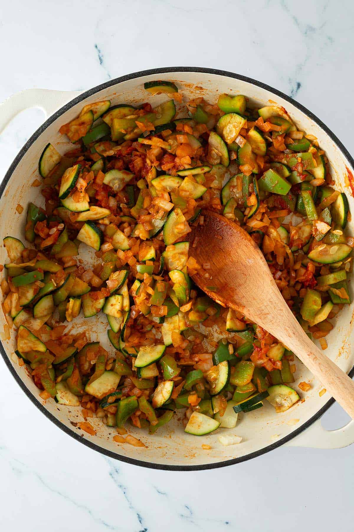 Pfanne mit angebratenem Gem&uuml;se aus Zucchini, Paprika, Zwiebeln, Gew&uuml;rzen und Tomatenmark das mit einem Holzl&ouml;ffel vermischt wird.