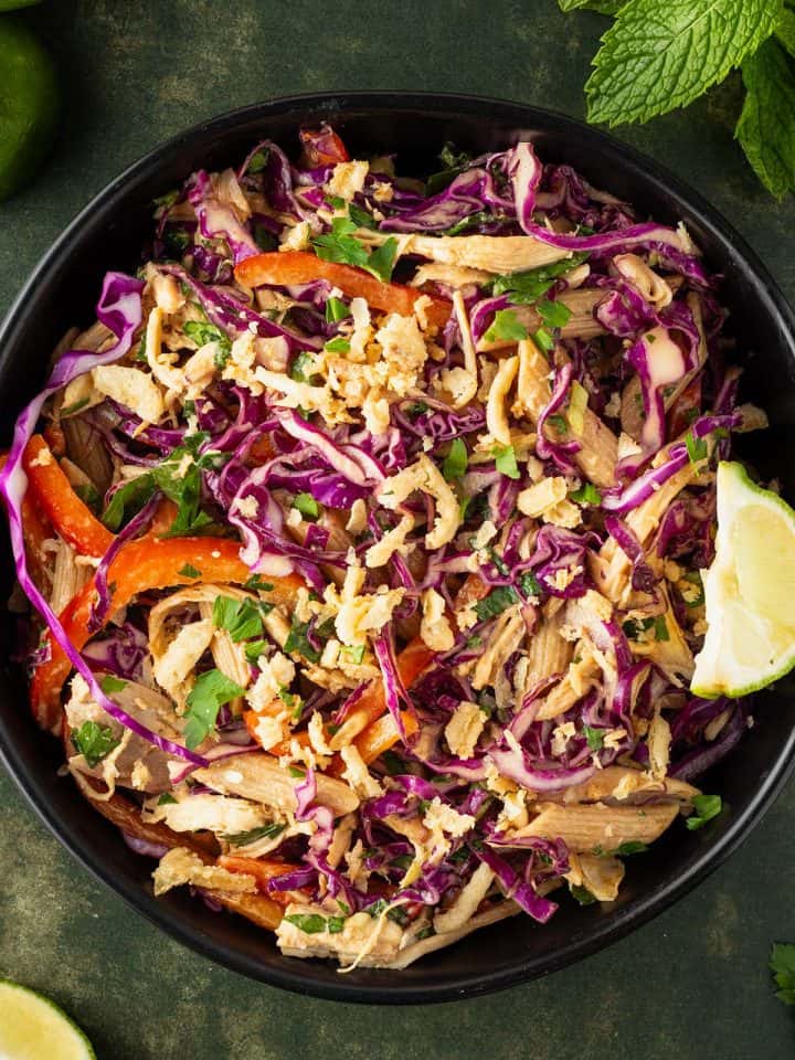 Ein Teller Thai Crunch Salat mit Rotkohl, Nudeln, Gem&uuml;se, H&auml;hnchen und R&ouml;stzwiebeln, Deko im Hintergrund.
