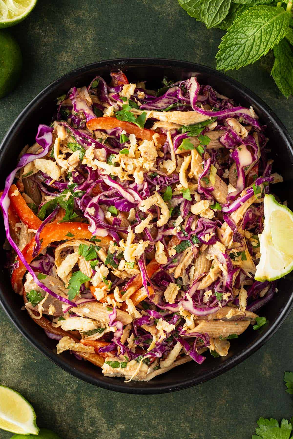 Ein Teller Thai Crunch Salat mit Rotkohl, Nudeln, Gem&uuml;se, H&auml;hnchen und R&ouml;stzwiebeln, Deko im Hintergrund.